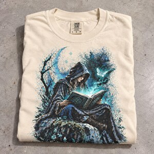 May include: A cream-colored short-sleeve t-shirt featuring a graphic print of a hooded figure sitting on a rock and reading a glowing book. The design includes a crescent moon, a moth, and blue foliage. The neck label reads Comfort Colors L G G.
