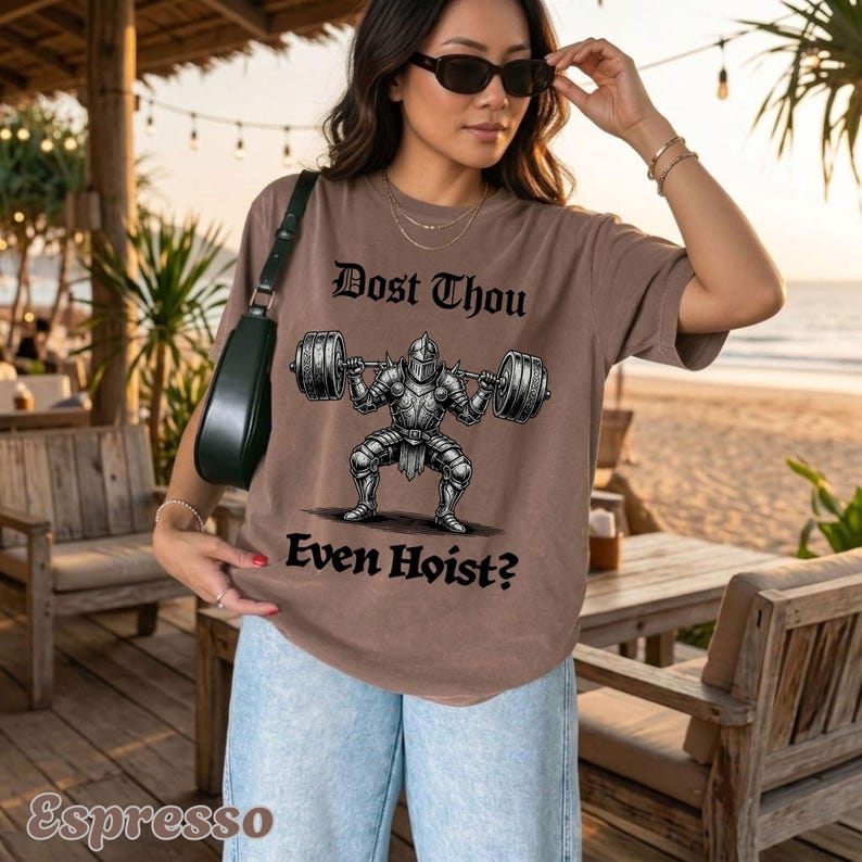 Op de afbeelding: Espresso-kleurig T-shirt met een afbeelding van een ridder die een halter optilt. De tekst "Dost Thou Even Hoist?" staat boven en onder de illustratie. Het shirt is een casual kledingstuk.