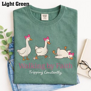 May include: Light green t-shirt with a graphic of three cartoon geese wearing pink bows. The text "Walking by Faith Tripping Constantly" is printed below the geese. The shirt is folded on top of blue jeans.