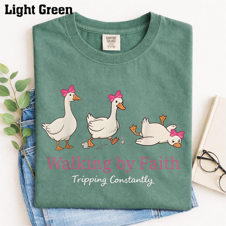 May include: Light green t-shirt with a graphic of three cartoon geese wearing pink bows. The text "Walking by Faith Tripping Constantly" is printed below the geese. The shirt is folded on top of blue jeans.