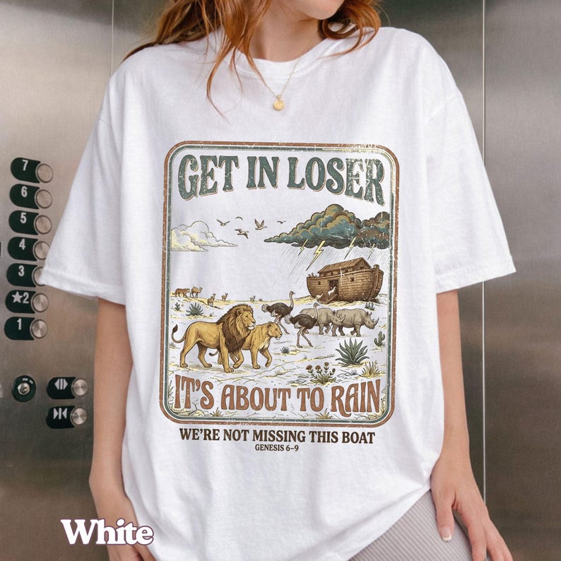 Op de afbeelding: Wit T-shirt met een grafisch ontwerp met de tekst "GET IN LOSER" boven een illustratie van dieren, een boot en regen. De zin "IT'S ABOUT TO RAIN" staat onder de illustratie, met de tekst "WE'RE NOT MISSING THIS BOAT" en "GENESIS 6-9" eronder.