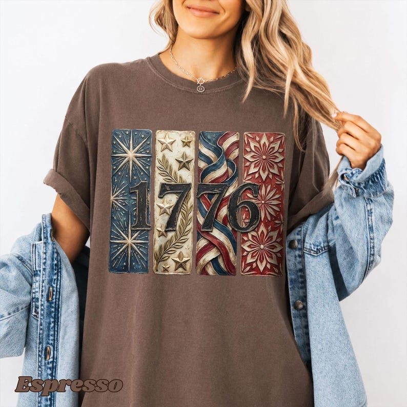 May include: Brown t-shirt with the number "1776" in a patriotic design. The design features stars, stripes, and floral patterns in red, white, and blue. The shirt has short sleeves and a relaxed fit.