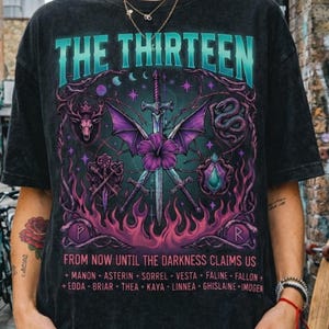 May include: Black t-shirt with a graphic design featuring the text "THE THIRTEEN" in teal and purple. The design includes swords, a deer head, a snake, and the text "FROM NOW UNTIL THE DARKNESS CLAIMS US".