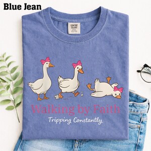 May include: A blue t-shirt with a graphic of three cartoon geese, each wearing a pink bow. The geese are in different poses, with one tripping. The text "Walking by Faith, Tripping Constantly" is printed below the geese.