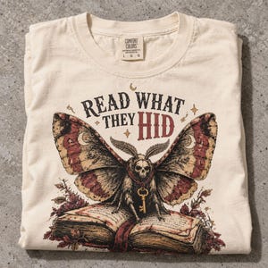 May include: A cream-colored t-shirt featuring a graphic of a moth with a skull head, wearing a gold key necklace, perched on an open book. The text above reads "READ WHAT THEY HID" in black and red lettering. The shirt has a Comfort Colors label.
