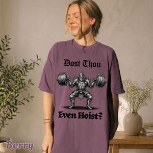 Op de afbeelding: Bessenkleurig T-shirt met de tekst "Dost Thou Even Hoist?" boven en onder een afbeelding van een ridder die gewichten heft. De ridder draagt een harnas en houdt een halter vast met gewichten aan beide uiteinden.