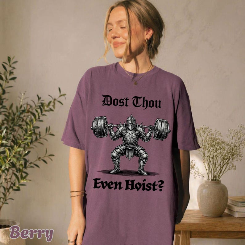 Op de afbeelding: Bessenkleurig T-shirt met de tekst "Dost Thou Even Hoist?" boven en onder een afbeelding van een ridder die gewichten heft. De ridder draagt een harnas en houdt een halter vast met gewichten aan beide uiteinden.