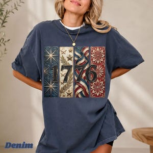 May include: A denim blue t-shirt featuring the number "1776" in a patriotic design. The design incorporates stars, stripes, and floral elements in red, white, and blue. The word "Denim" is printed at the bottom of the shirt.