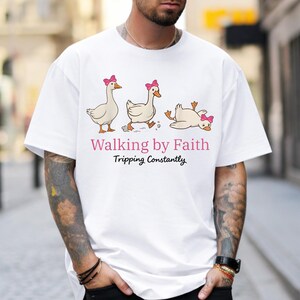May include: White t-shirt featuring a cartoon illustration of three geese with pink bows. The geese are in different poses, with one tripping. The text "Walking by Faith Tripping Constantly" is printed below the geese.