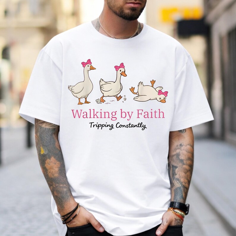 May include: White t-shirt featuring a cartoon illustration of three geese with pink bows. The geese are in different poses, with one tripping. The text "Walking by Faith Tripping Constantly" is printed below the geese.