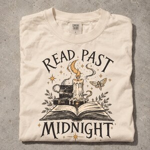 May include: A cream-colored t-shirt featuring a graphic print of an open book, a stack of books, a lit candle, a crescent moon, and a moth. The text "READ PAST MIDNIGHT" is printed in black lettering above and below the central illustration.