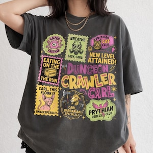 May include: Dark gray t-shirt with a colorful graphic design. The design includes various stamps with text like "GAZER FRIEND!", "EATING ON THE RUN", and "DUNGEON CRAWLER CARL". Other stamps feature a cat, a gas mask, and a goat.