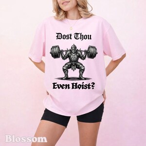 Op de afbeelding: Lichtroze t-shirt met de tekst "Dost Thou Even Hoist?" boven en onder een afbeelding van een ridder die gewichten heft. De ridder draagt een harnas en tilt een halterstang op.
