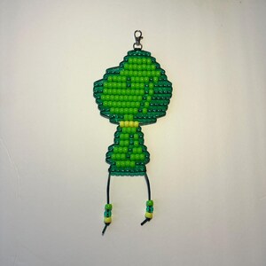 May include: A beaded keychain crafted from small, vibrant green and lime-colored plastic beads arranged in a stylized character shape. The figure features a large head and a smaller body, topped with a silver-toned metal lobster claw clasp.