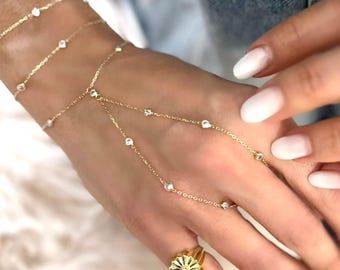 May include: Gold-toned jewelry set featuring a hand chain, bracelet, and ring. The hand chain connects to the ring with delicate chains adorned with small, clear gemstones. The bracelet and ring also feature similar gemstones. The ring has a floral design.