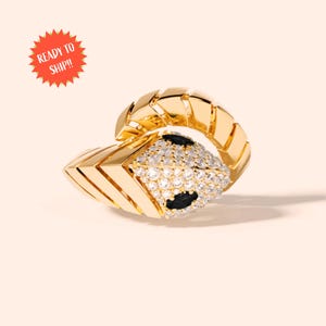May include: A gold-toned ring in a snake design, featuring a head encrusted with clear and black gemstones. The ring has a geometric pattern and is set against a soft, neutral background. The ring is ready to ship.