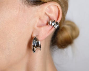 May include: A pair of silver hoop earrings and a matching ear cuff. The earrings have a unique, asymmetrical design. The ear cuff features a ridged, geometric pattern. The jewelry is worn on a person's ear.