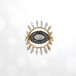 May include: A gold ring featuring a black and white diamond eye design. The eye is encircled by a ring of smaller diamonds.