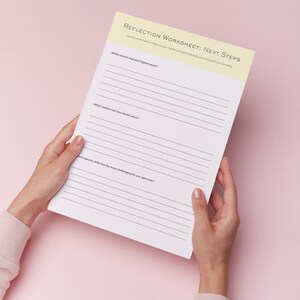 以下が含まれることがあります： ピンク色の背景に、手が持っている「Reflection Worksheet: Next Steps」というタイトルの白いワークシート。ワークシートの上部には、テキストが書かれた黄色のボーダーがあります。