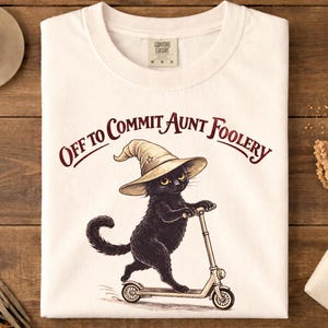 May include: White t-shirt with the phrase "Off to Commit Aunt Foolery" in a burgundy font. The graphic features a black cat wearing a witch hat and riding a scooter. The shirt is folded on a wooden surface.