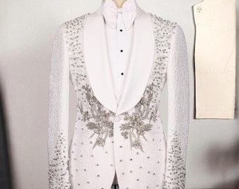 May include: A white tuxedo jacket with a shawl collar, adorned with silver embellishments and floral embroidery. The jacket is displayed on a mannequin, paired with a white dress shirt and bow tie. A pattern piece is visible in the background.