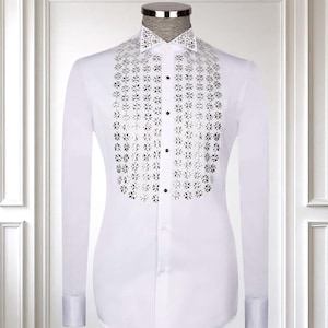 May include: A white dress shirt with long sleeves and a spread collar. The shirt features a decorative, symmetrical design of silver-colored cutouts down the front. The cuffs are buttoned, and the shirt is displayed on a mannequin.