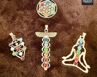Può includere: Quattro ciondoli color argento con accenti di pietre preziose colorate. I disegni includono un fiore della vita, un albero della vita, un totem e una figura meditante. I ciondoli sono esposti su una superficie marrone strutturata.