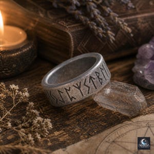 May include: A silver-colored ring with engraved runic symbols. The ring is displayed on a wooden surface with a crystal, dried flowers, and a lit candle in the background. The ring's design suggests a theme of Norse mythology or ancient symbolism.