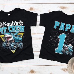 以下が含まれることがあります： 2つの黒いTシャツ。モンスター・トラックのデザインで、1つは「Noah's PIT CREW」とサメをテーマにしたトラックが描かれています。もう1つは「PAPA 1」と似たようなトラックのデザインです。どちらのシャツも明るい木製の表面にあります。