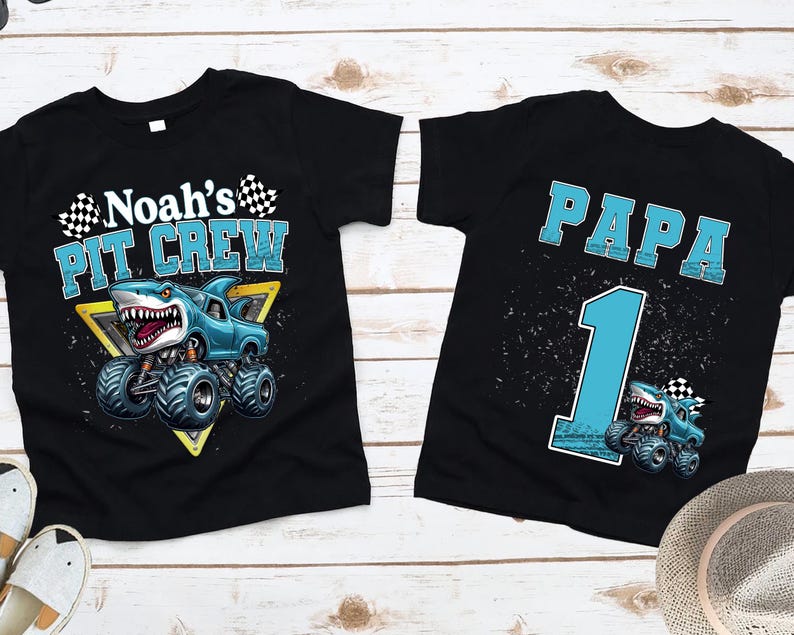 以下が含まれることがあります： 2つの黒いTシャツ。モンスター・トラックのデザインで、1つは「Noah's PIT CREW」とサメをテーマにしたトラックが描かれています。もう1つは「PAPA 1」と似たようなトラックのデザインです。どちらのシャツも明るい木製の表面にあります。