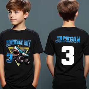 May include: Black short-sleeved t-shirt with a shark-themed graphic. The front features the text "BIRTHDAY BOY" and the number "3" with a shark on a monster truck. The back displays the name "JACKSON" and the number "3".