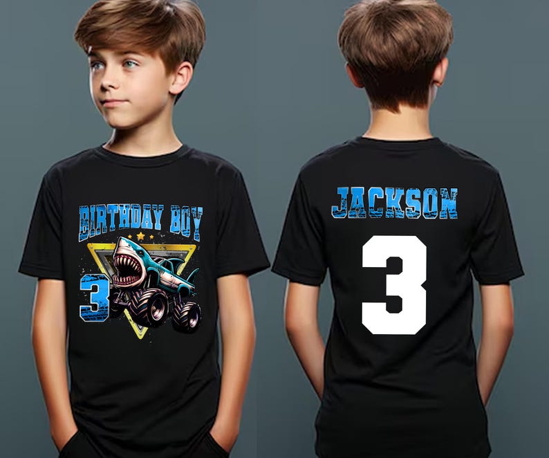 May include: Black short-sleeved t-shirt with a shark-themed graphic. The front features the text "BIRTHDAY BOY" and the number "3" with a shark on a monster truck. The back displays the name "JACKSON" and the number "3".