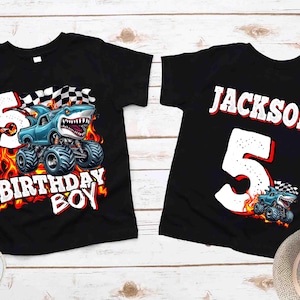 Puede incluir: Dos camisetas negras con un dise&ntilde;o de monster truck. Una camiseta dice "BIRTHDAY BOY" con un cami&oacute;n con tem&aacute;tica de tibur&oacute;n y el n&uacute;mero 5. La otra camiseta tiene el nombre "JACKSON" y el n&uacute;mero 5, con un gr&aacute;fico de cami&oacute;n m&aacute;s peque&ntilde;o.