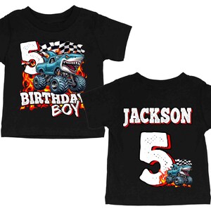 Puede incluir: Dos camisetas negras para ni&ntilde;os peque&ntilde;os. Una presenta un cami&oacute;n monstruo azul con un dise&ntilde;o de tibur&oacute;n, el n&uacute;mero 5 y el texto "BIRTHDAY BOY". La otra camiseta tiene el nombre "JACKSON", el n&uacute;mero 5 y un gr&aacute;fico de cami&oacute;n monstruo.