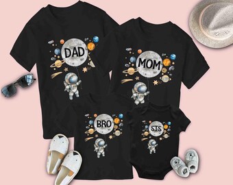 Puede incluir: Un conjunto de cuatro camisetas negras con temática espacial. Las camisetas presentan una luna con planetas y estrellas, y las palabras "Papá", "Mamá", "Hermano" y "Hermana" en cada camiseta. Cada camiseta también presenta un astronauta de dibujos animados.