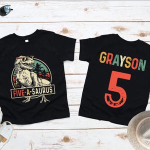 May include: Two black t-shirts. One features a dinosaur wearing sunglasses with the text "FIVE-A-SAURUS". The other shirt has the name "GRAYSON" and the number "5" in a colorful, retro font. Sunglasses, sandals, and a hat are also in the image.