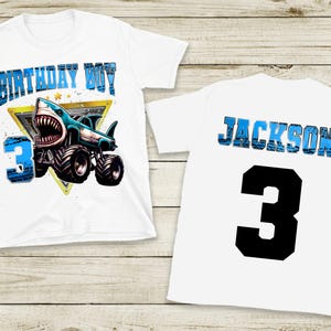 May include: White t-shirt featuring a shark monster truck design with the text "BIRTHDAY BOY" and the number "3". The back of the shirt displays the name "JACKSON" and the number "3".