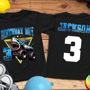 May include: Black t-shirts with a shark-themed design. One shirt features a shark monster truck with the text "BIRTHDAY BOY" and the number "3". The other shirt has the name "JACKSON" and the number "3".