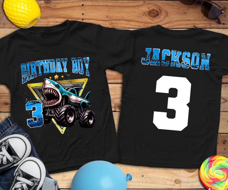 May include: Black t-shirts with a shark-themed design. One shirt features a shark monster truck with the text "BIRTHDAY BOY" and the number "3". The other shirt has the name "JACKSON" and the number "3".