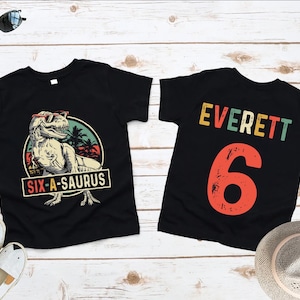 May include: Two black t-shirts. One features a dinosaur wearing sunglasses with the text "SIX-A-SAURUS". The other shirt has the name "Everett" and the number "6" in a retro font. Both shirts are on a light wood surface.