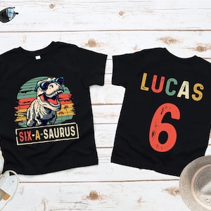 May include: Two black t-shirts with a dinosaur graphic and the text "SIX-A-SAURUS" and "LUCAS 6". The dinosaur wears sunglasses against a sunset background. The second shirt has the name "LUCAS" and the number "6" in colorful letters.