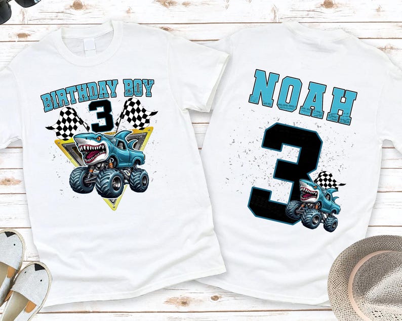 以下が含まれることがあります： サメをテーマにしたモンスターカーのグラフィックがプリントされた白いTシャツ。「BIRTHDAY BOY 3」とチェック柄の旗が前面にプリントされています。Tシャツの背面には「NOAH」という名前と数字の「3」がプリントされています。