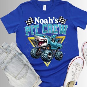 以下が含まれることがあります： 「Noah's Pit Crew」の文字とサメをテーマにしたモンスターカーのグラフィックが特徴のロイヤルブルーのTシャツ。デザインには、チェック柄の旗と黄色の三角形が含まれています。このシャツは、デニムショーツと白いスニーカーと組み合わせています。