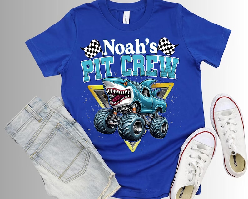以下が含まれることがあります： 「Noah's Pit Crew」の文字とサメをテーマにしたモンスターカーのグラフィックが特徴のロイヤルブルーのTシャツ。デザインには、チェック柄の旗と黄色の三角形が含まれています。このシャツは、デニムショーツと白いスニーカーと組み合わせています。