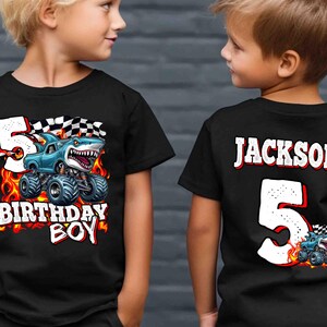 Puede incluir: Camiseta negra con un dise&ntilde;o de cami&oacute;n monstruo con una cabeza de tibur&oacute;n, llamas y el texto "5 BIRTHAY BOY". La parte trasera de la camiseta tiene el nombre "JACKSON" y el n&uacute;mero "5".