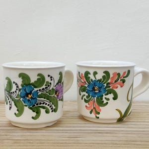 May include: Two white ceramic mugs with floral designs. One mug has a blue, pink, and green floral design. The other mug has a blue, green, and orange floral design.