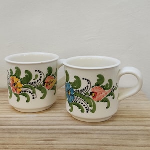 May include: Two white ceramic mugs with floral designs. The mugs have green leaves, black swirls, and flowers in shades of yellow, orange, pink, and blue.