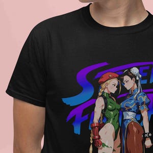 May include: Black t-shirt featuring a graphic of two female characters from the Street Fighter video game. One character wears a green bodysuit and the other a blue dress. The shirt has the Street Fighter logo in blue and purple.