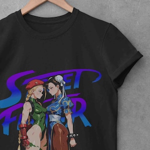 May include: Black t-shirt featuring a graphic of two female characters from the Street Fighter video game. One character wears a green bodysuit and the other a blue dress. The text "Street Fighter" is in purple and blue.