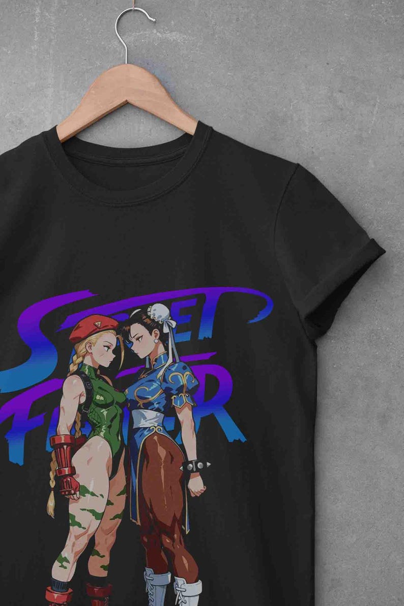 May include: Black t-shirt featuring a graphic of two female characters from the Street Fighter video game. One character wears a green bodysuit and the other a blue dress. The text "Street Fighter" is in purple and blue.